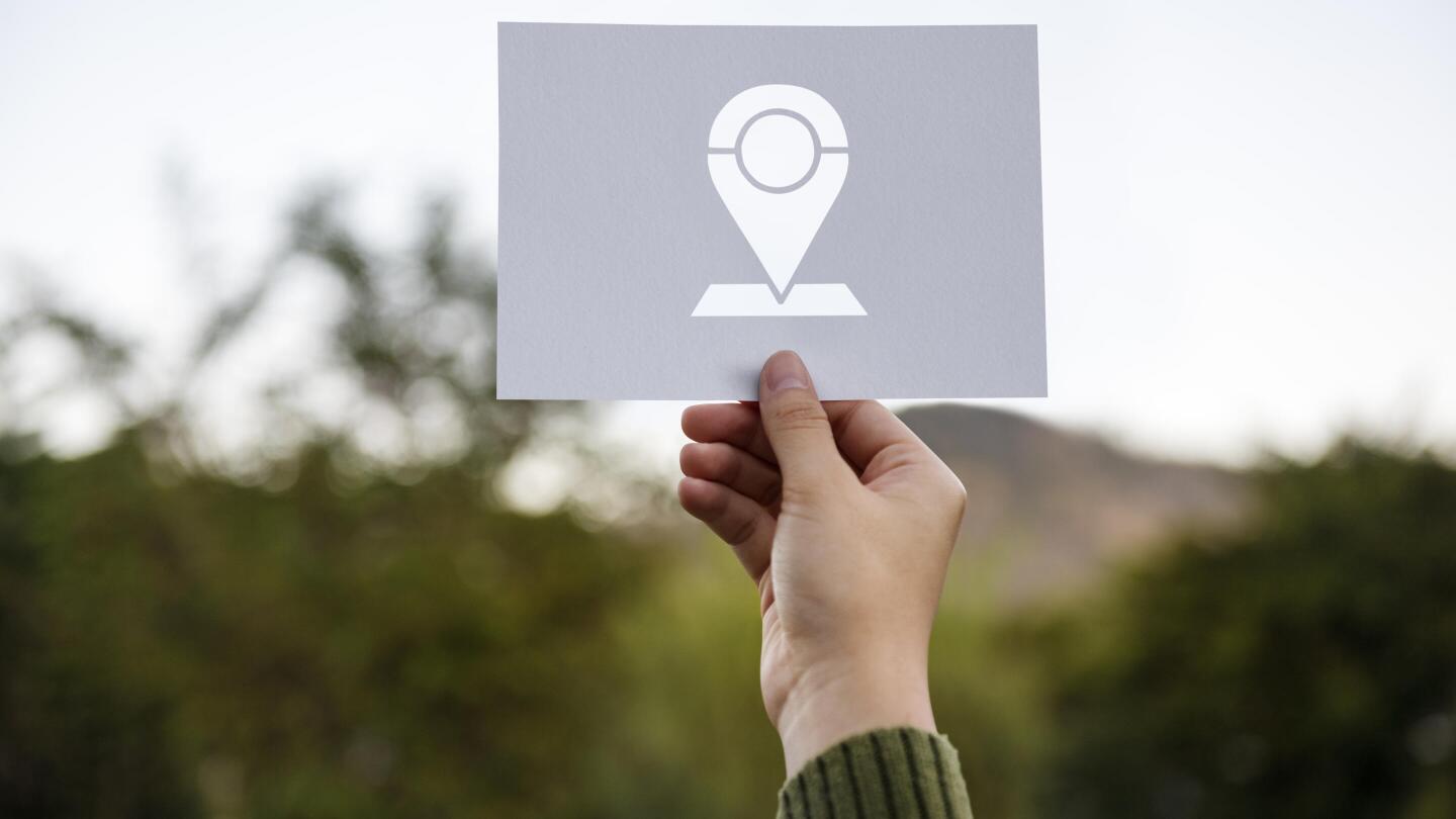 A hand holds up a sign with a location pin icon against a blurry natural landscape.