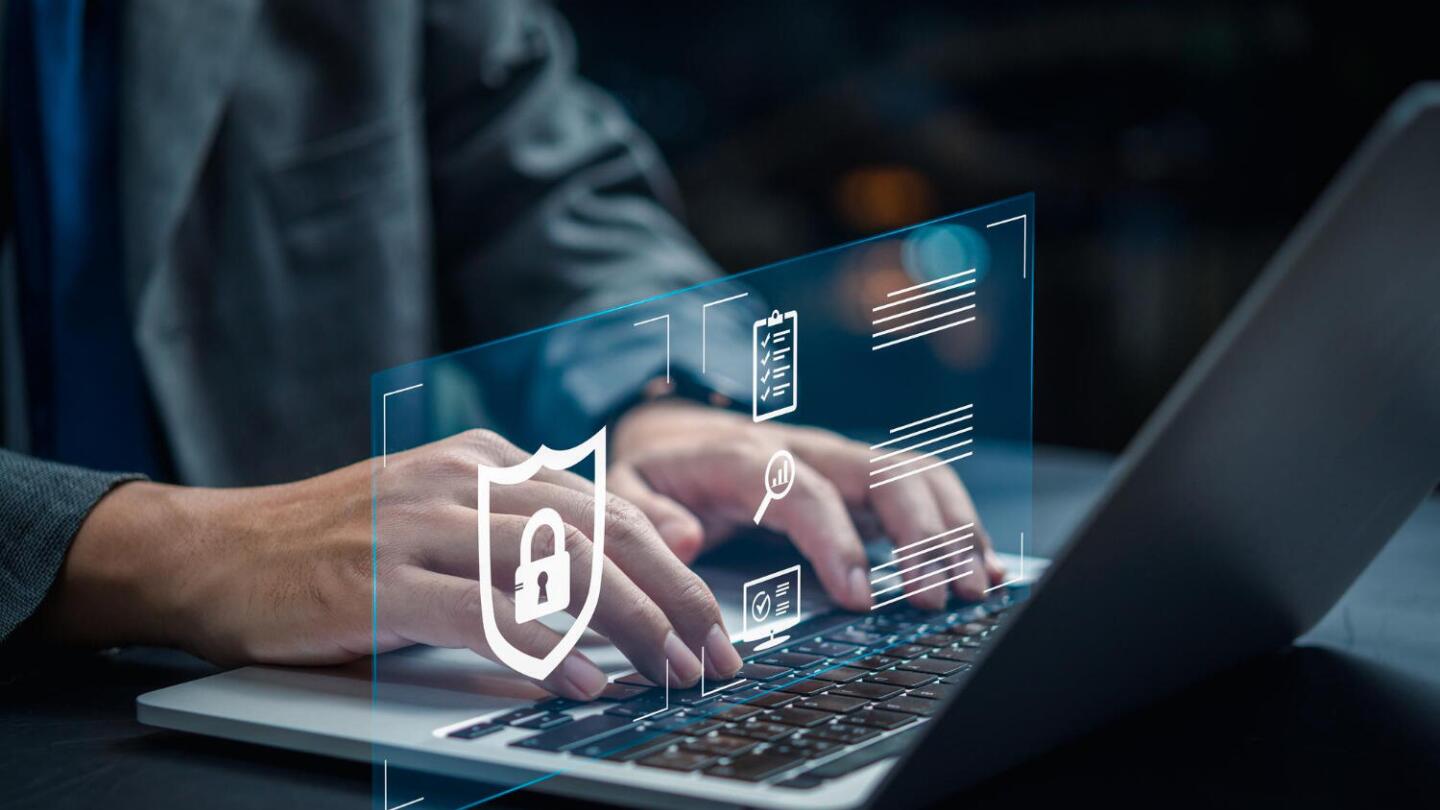 Ein Mann im Anzug tippt auf einem Laptop, über dem eine transparente Grafik mit Symbolen zur Cybersicherheit, wie einem Schild mit Schloss, schwebt.