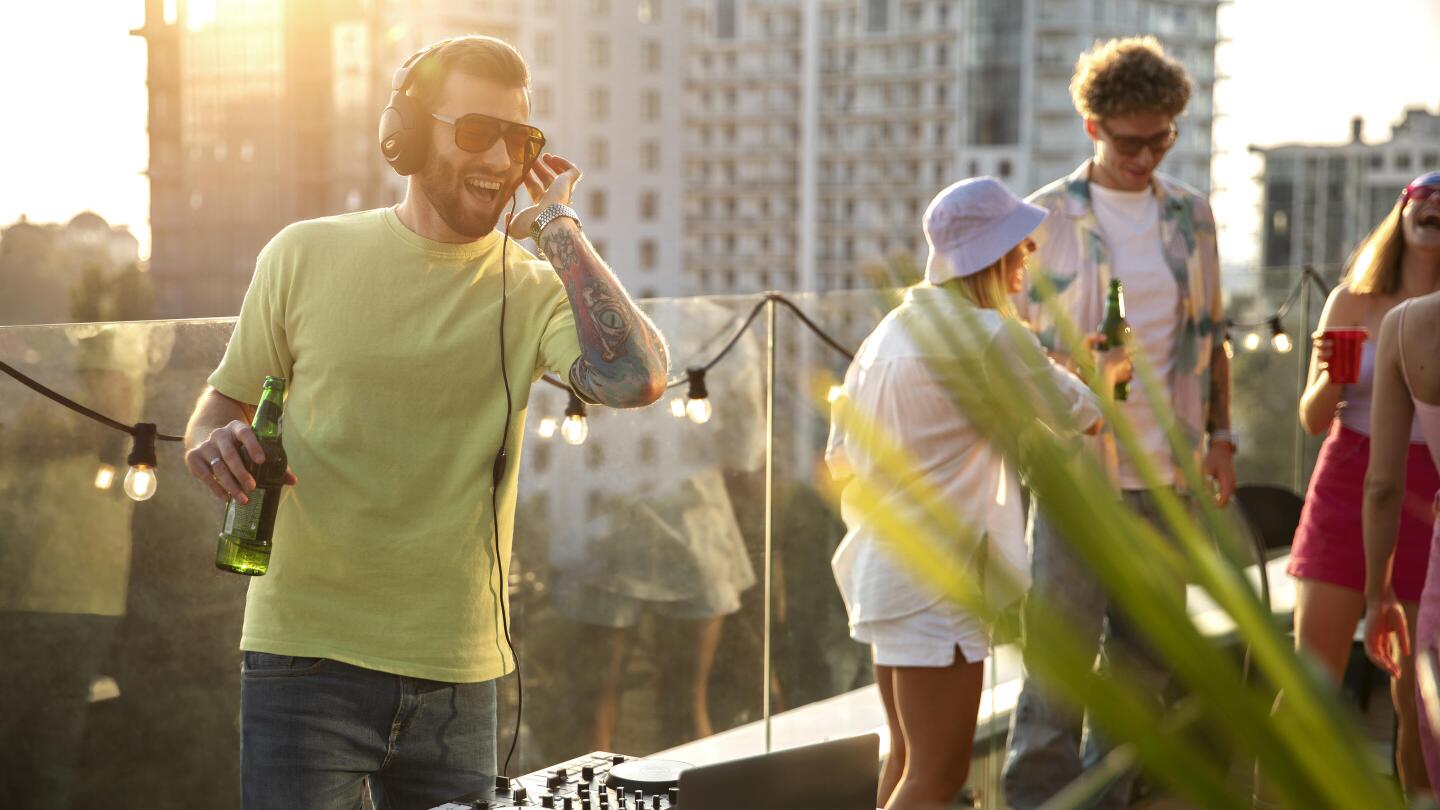 A DJ with headphones is playing a set at a rooftop party during sunset, while friends dance and talk around him.